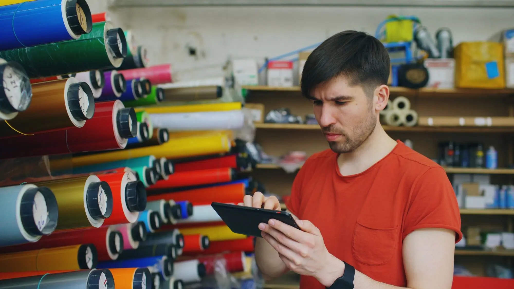 Print shop owner checking a renewal bill on a tablet beside racks of vinyl rolls in his workshop